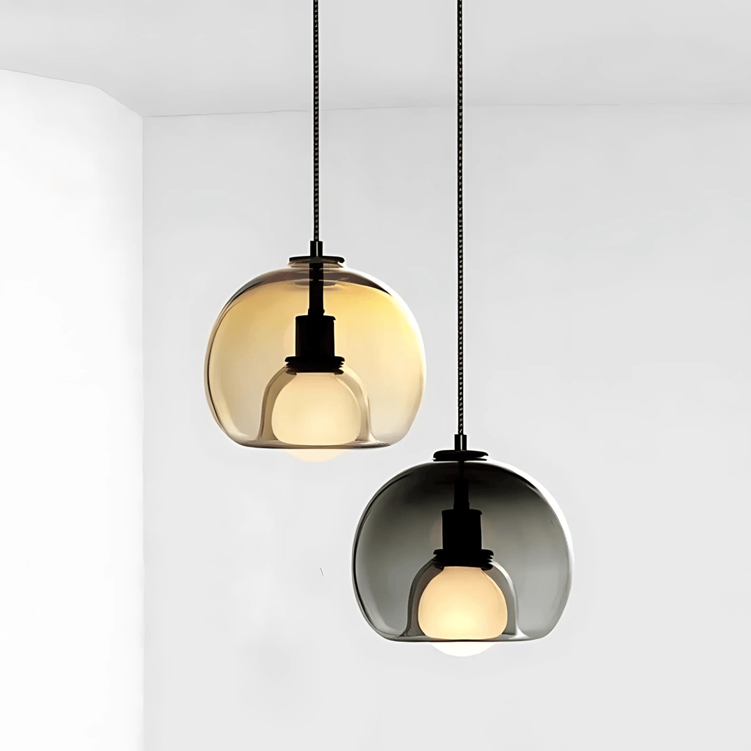 Two handblown glass pendant lights, one amber and one smoked grey, hanging against a white wall.