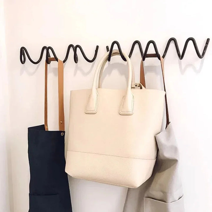 Wrought iron ocean wave wall hook rack with a cream tote bag, navy apron, and grey apron hanging.