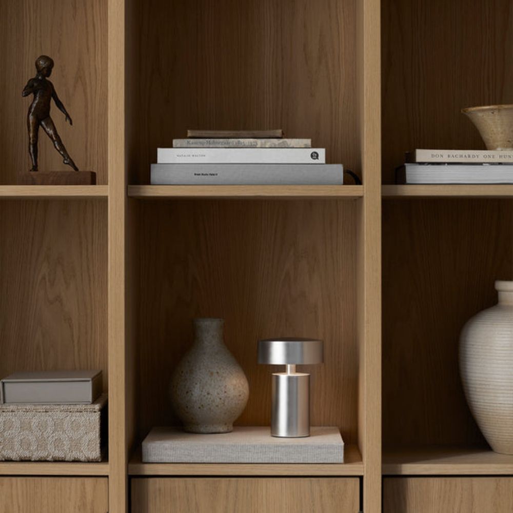 A silver column LED table lamp sits on a shelf next to a speckled vase.
