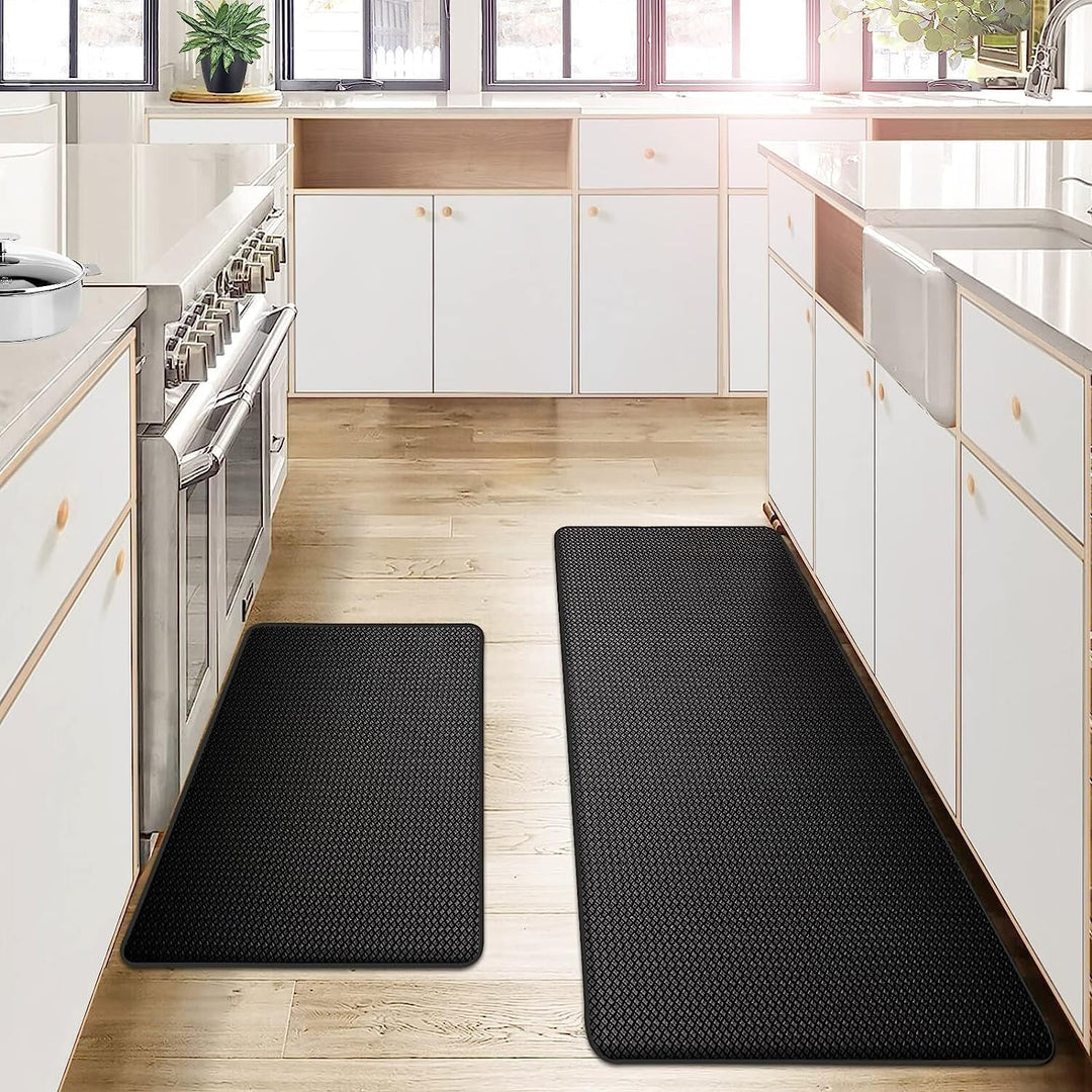 Two black anti-fatigue kitchen mats on a wooden floor in front of white cabinets and a stainless steel stove.