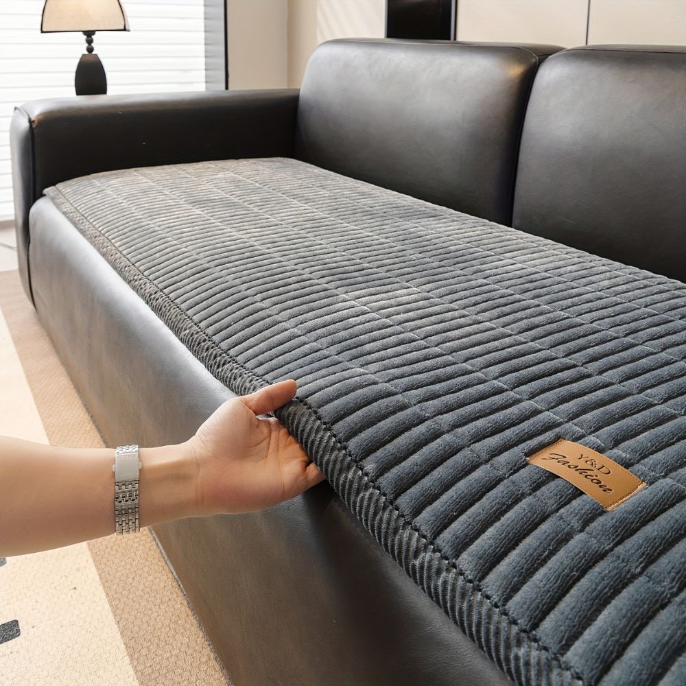A hand lifts the edge of a gray quilted couch cushion protector on a black leather sofa.