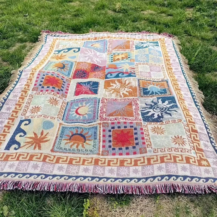 Outdoor picnic blanket with a colorful patchwork design of suns, waves, and geometric borders, laid on grass.