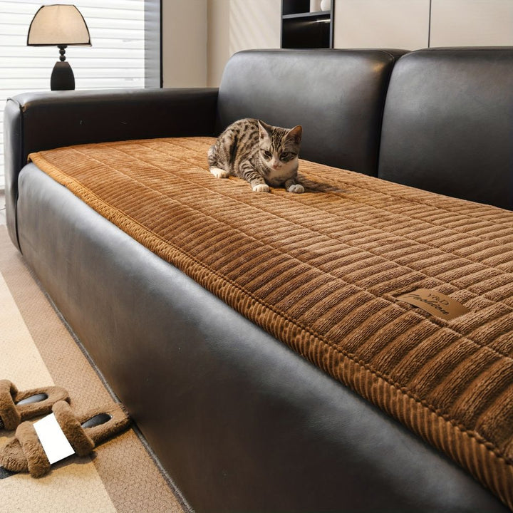 A tabby cat rests on a brown microfiber couch cover protector on a dark leather sofa.