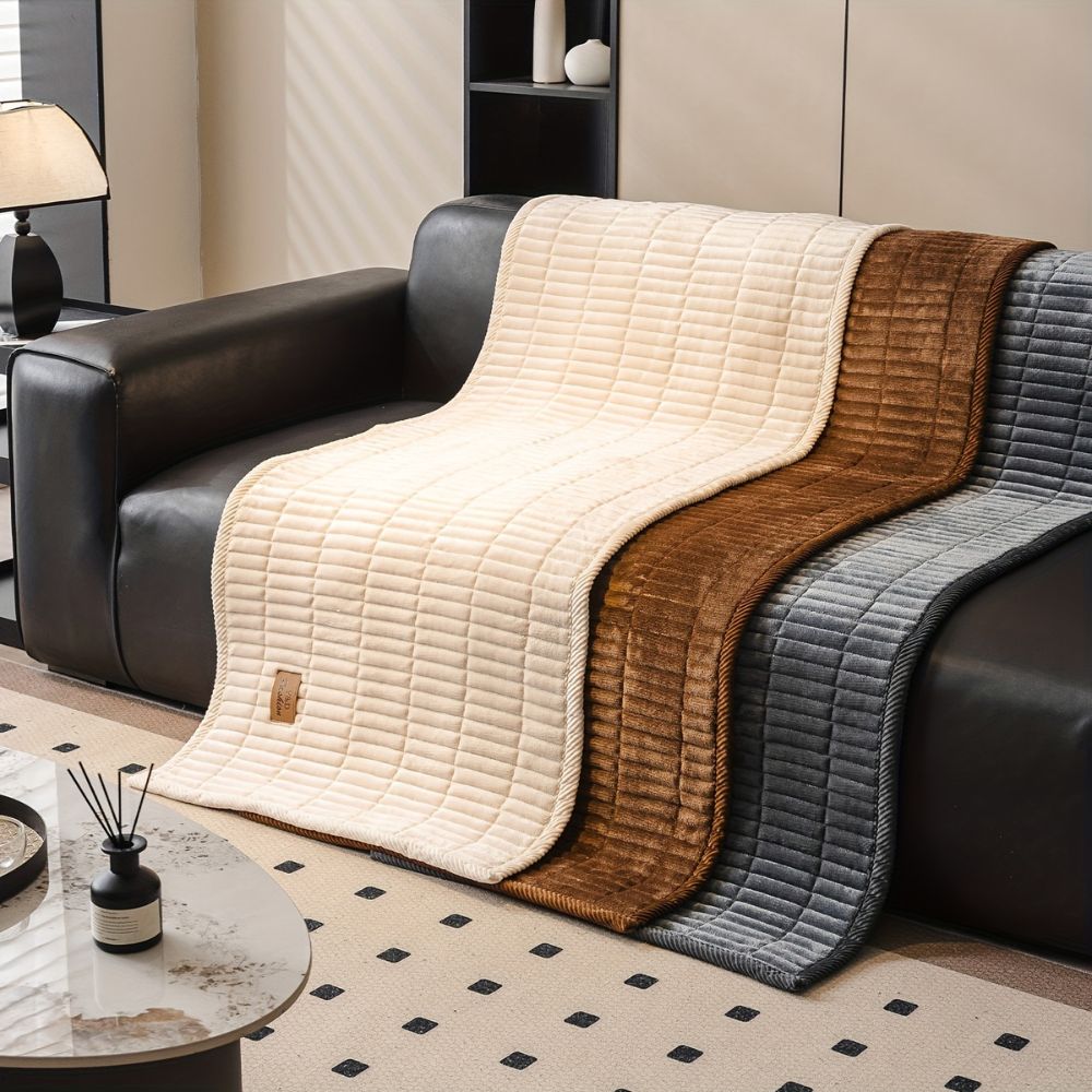 Three quilted microfiber couch covers in cream, brown, and gray draped over a dark leather sofa.