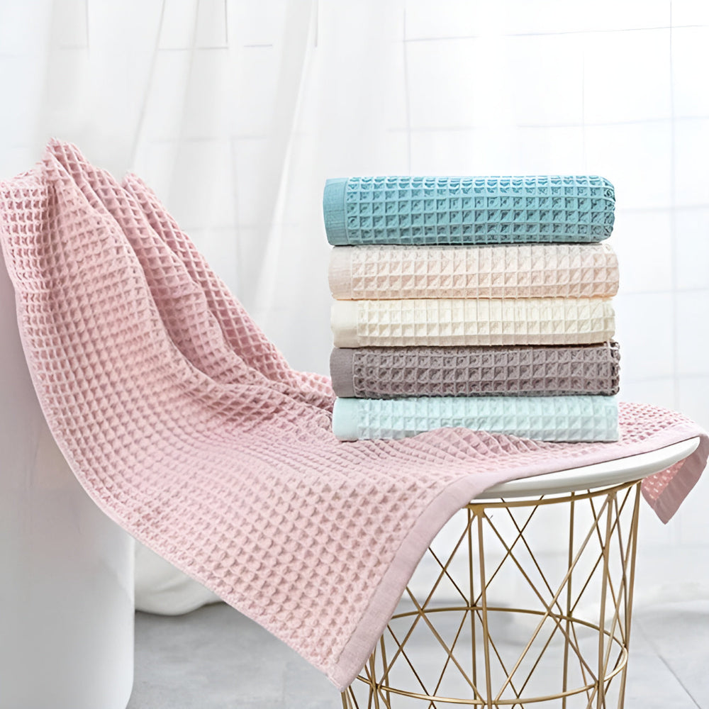 Stack of six waffle weave cotton towels in blue, beige, cream, grey, and light blue, with a pink towel draped over a white surface.