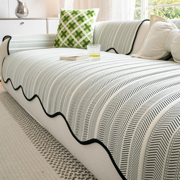 White and black herringbone sofa cover with a scalloped edge, draped over a sofa with a green patterned pillow and a glass of water.