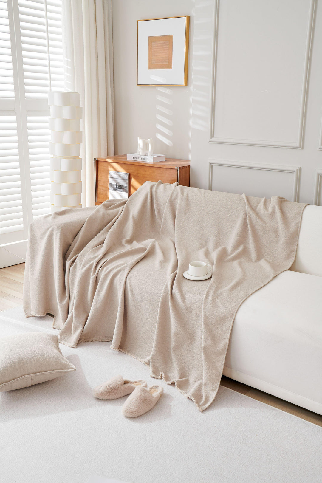 A milk tea colored soft throw blanket draped over a white sofa with a pillow and slippers on the floor.