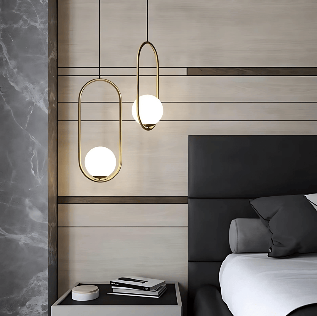 Two gold oval pendant lights with white glass globes hang over a nightstand next to a bed.