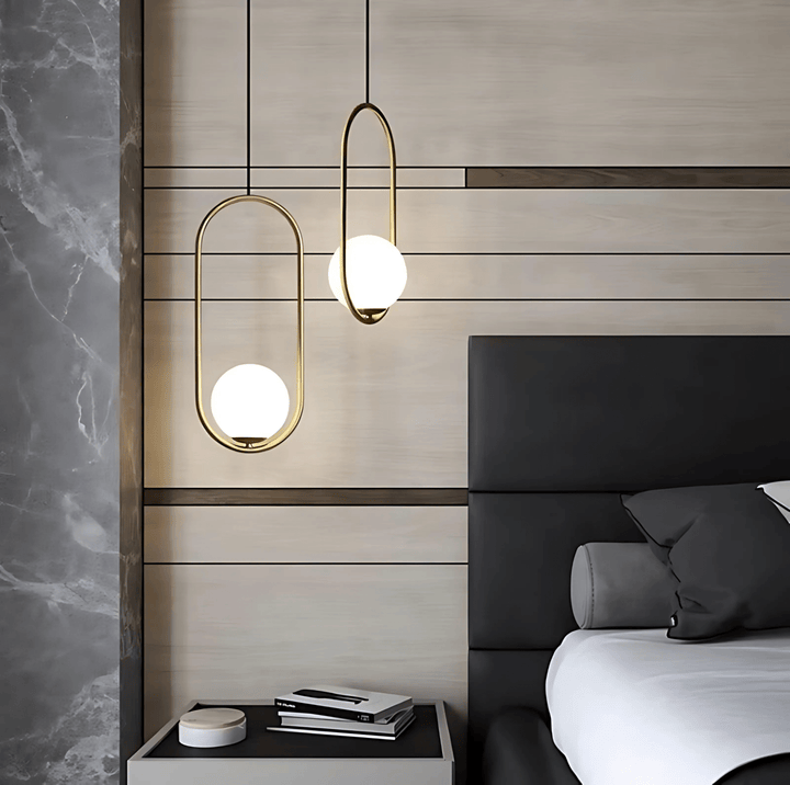 Two gold oval pendant lights with white glass globes hang over a nightstand next to a bed.
