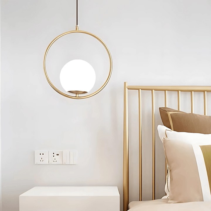 Gold ring pendant light with a white globe hangs above a white nightstand next to a bed.