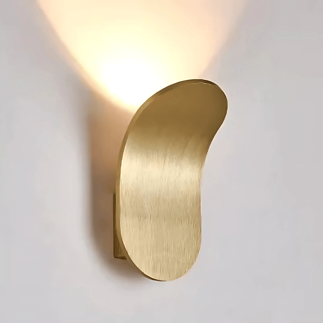 Brushed brass metal wall sconce with a curved, organic shape, casting a warm light upwards.