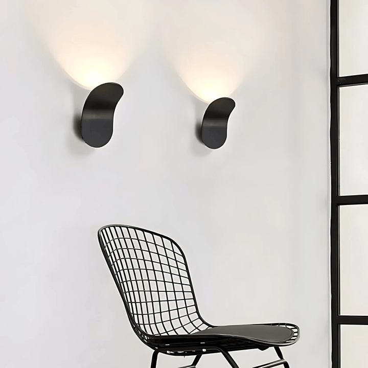 Two modern black metal wall sconces casting warm light on a white wall above a black wire chair.