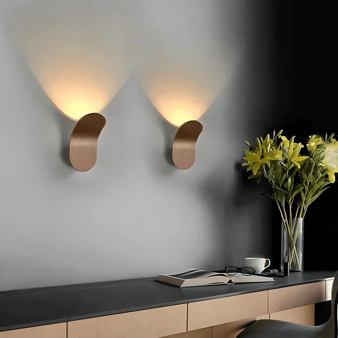 Two modern metal wall sconces with curved shapes cast warm light on a gray wall above a desk.