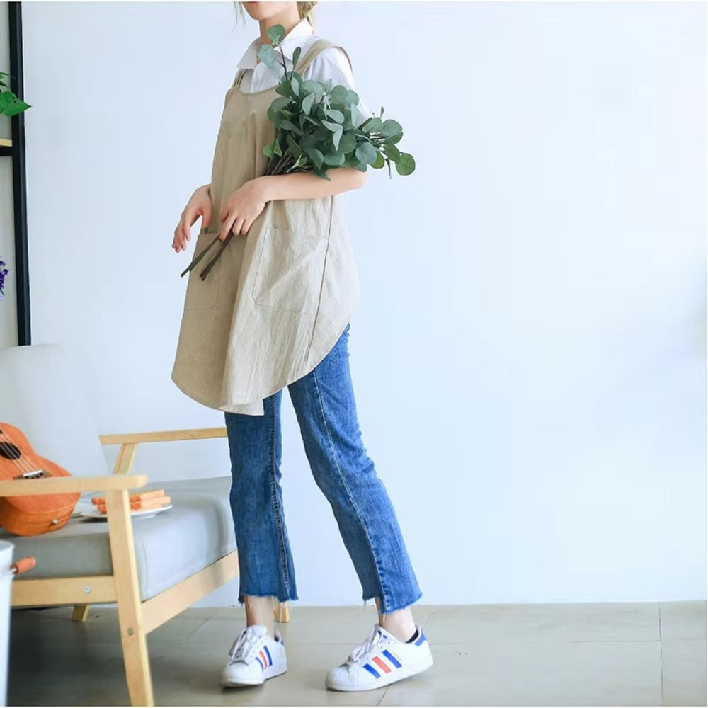 Woman wearing a beige linen cross-back apron over a white shirt and blue jeans, holding green eucalyptus branches.