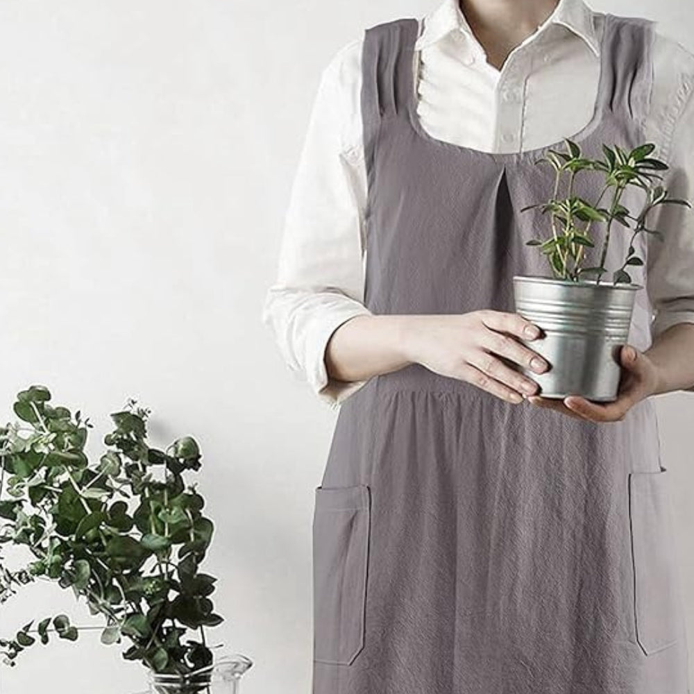 Person wearing a grey cross-back linen apron and holding a small potted plant.