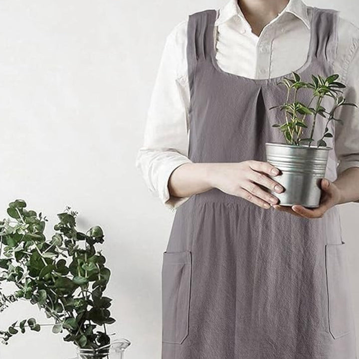 Person wearing a grey cross-back linen apron and holding a small potted plant.