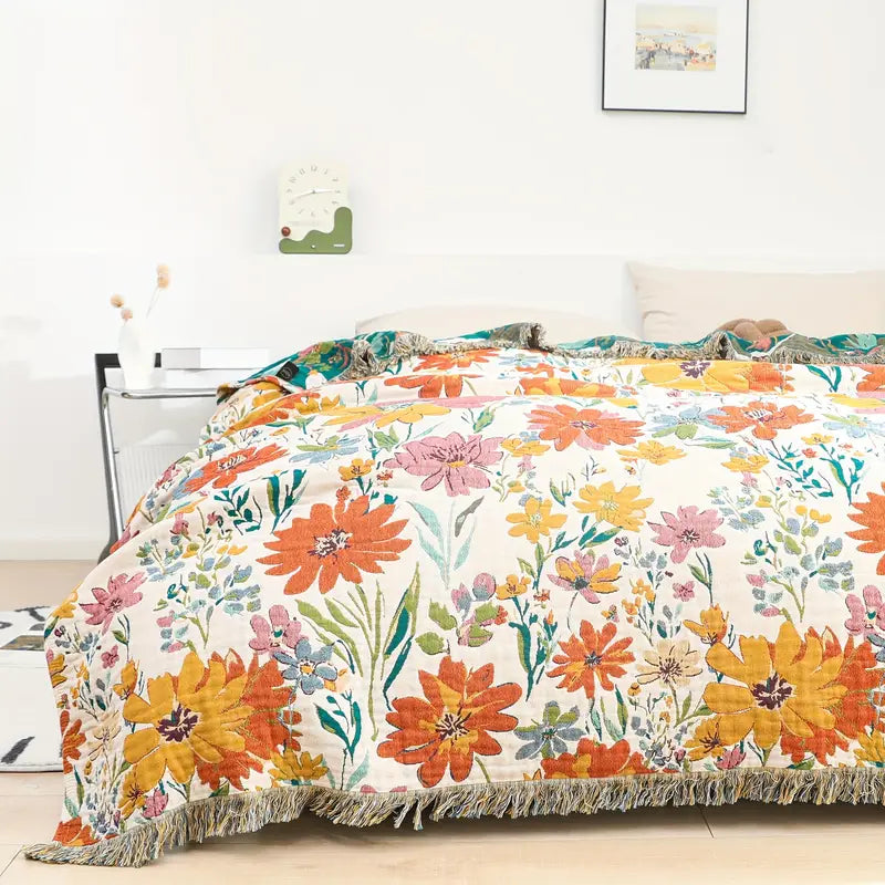 A cotton blanket with a vibrant, all-over floral print in shades of orange, yellow, pink, and blue on a cream background, with fringed edges.