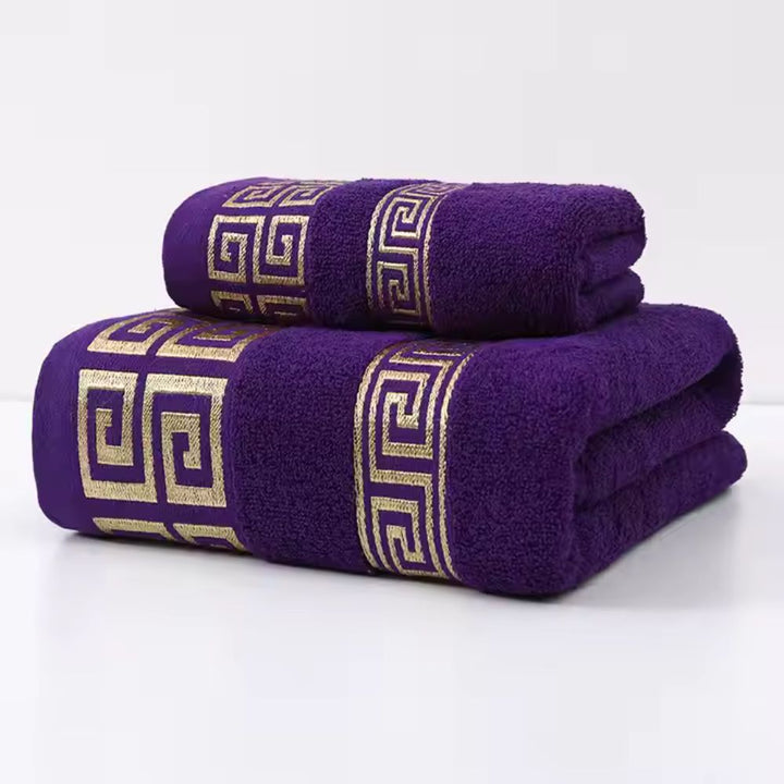 Two purple cotton towels with a gold Greek key pattern stacked on a white surface.
