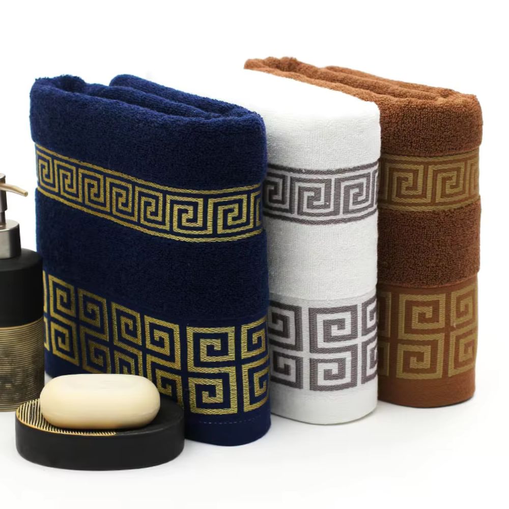 Three folded cotton towels: navy blue, white, and brown, each with a gold Greek key pattern.