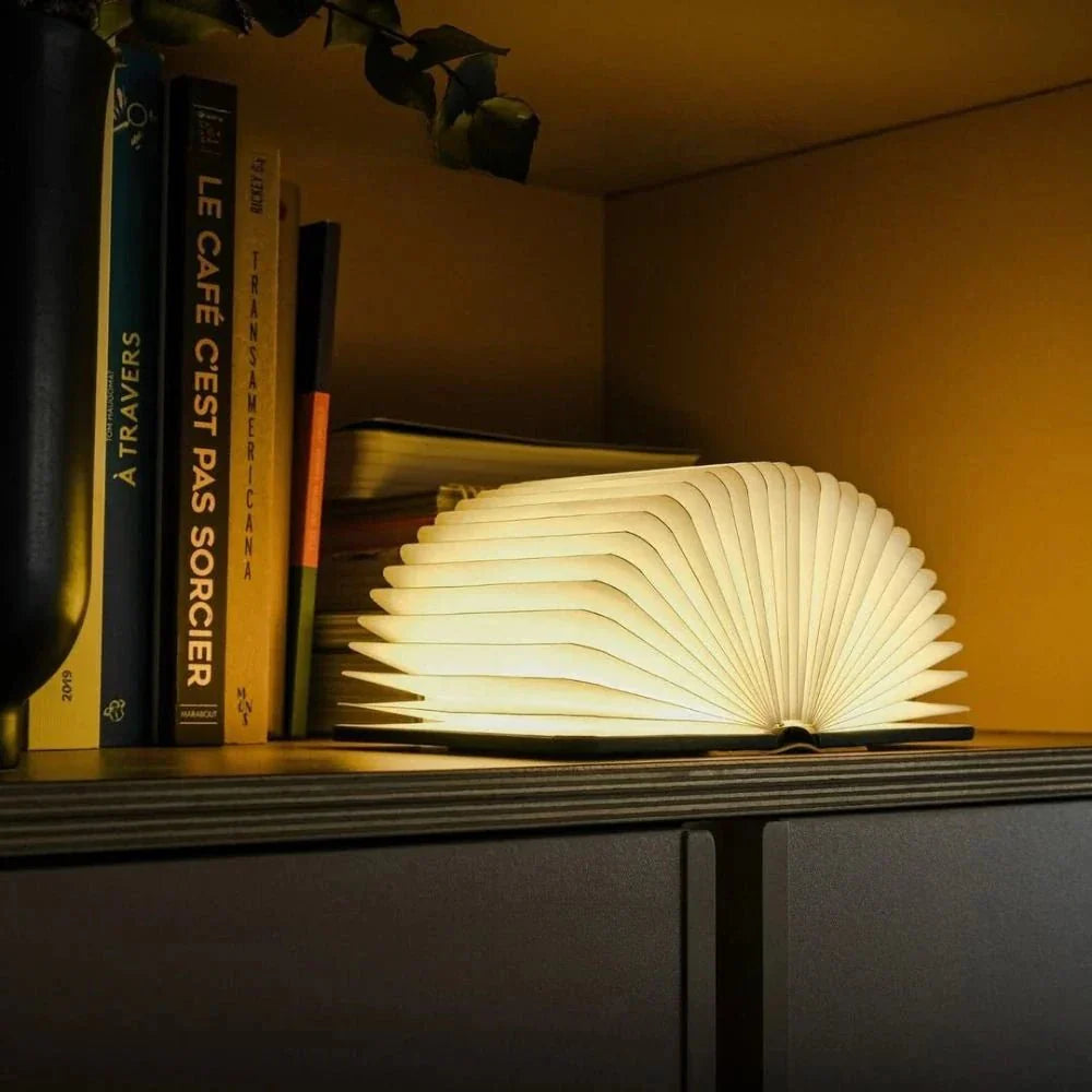 Foldable book lamp emitting warm light, placed on a shelf next to books.
