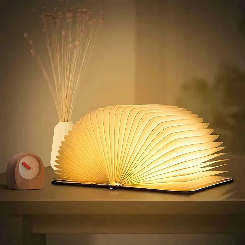 Foldable book lamp emitting warm light, placed on a table next to a vase of dried flowers and a small clock.