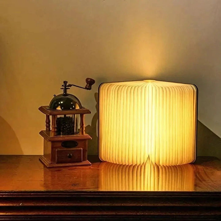 A wooden book lamp emitting warm light sits next to a vintage coffee grinder on a wooden table.