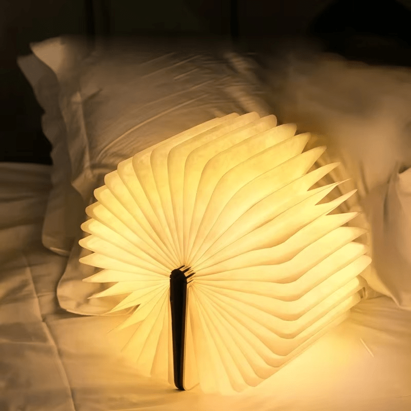 A book-shaped lamp is open and illuminated, casting a warm glow on a bed with white linens and pillows.