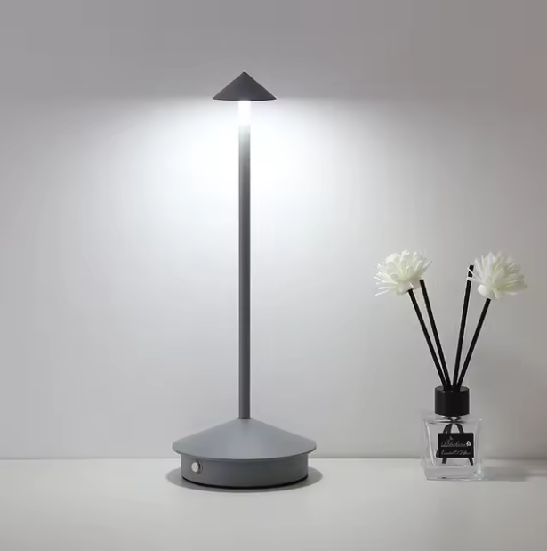 Gray cordless LED table lamp with a conical shade and base, illuminating a white wall.