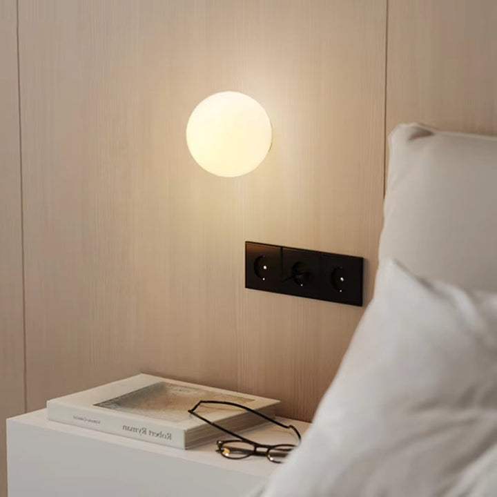Round glass wall sconce emitting warm light above a bedside table with a book and glasses.