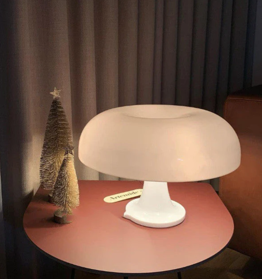 White mushroom-shaped table lamp with a soft glow on a pink table, next to two small decorative Christmas trees.