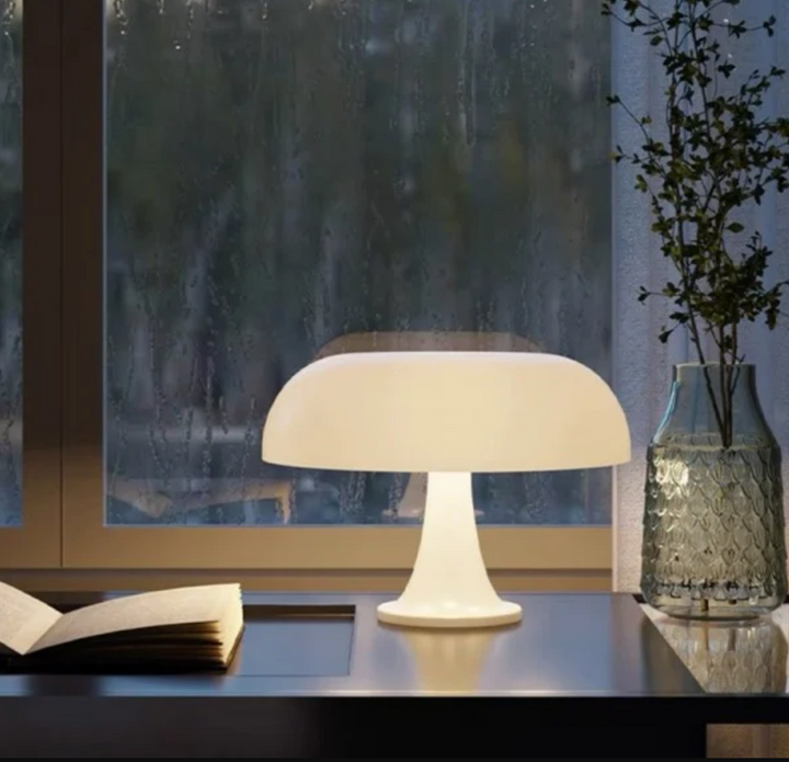 White mushroom-shaped LED table lamp on a desk next to an open book and a vase with greenery.