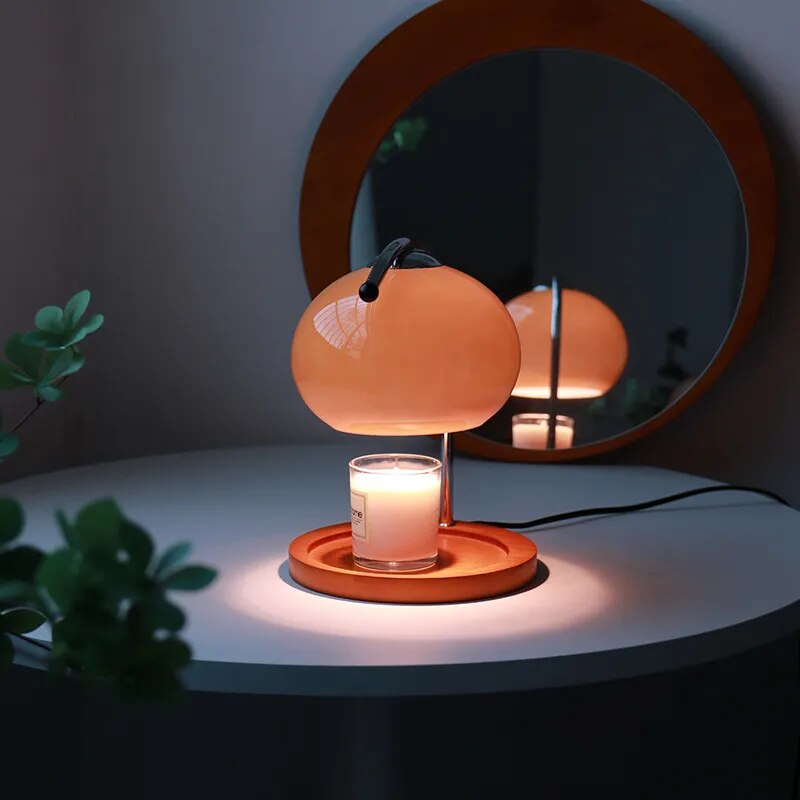 Orange glass candle warmer lamp with a lit candle on a wooden base, reflected in a mirror.