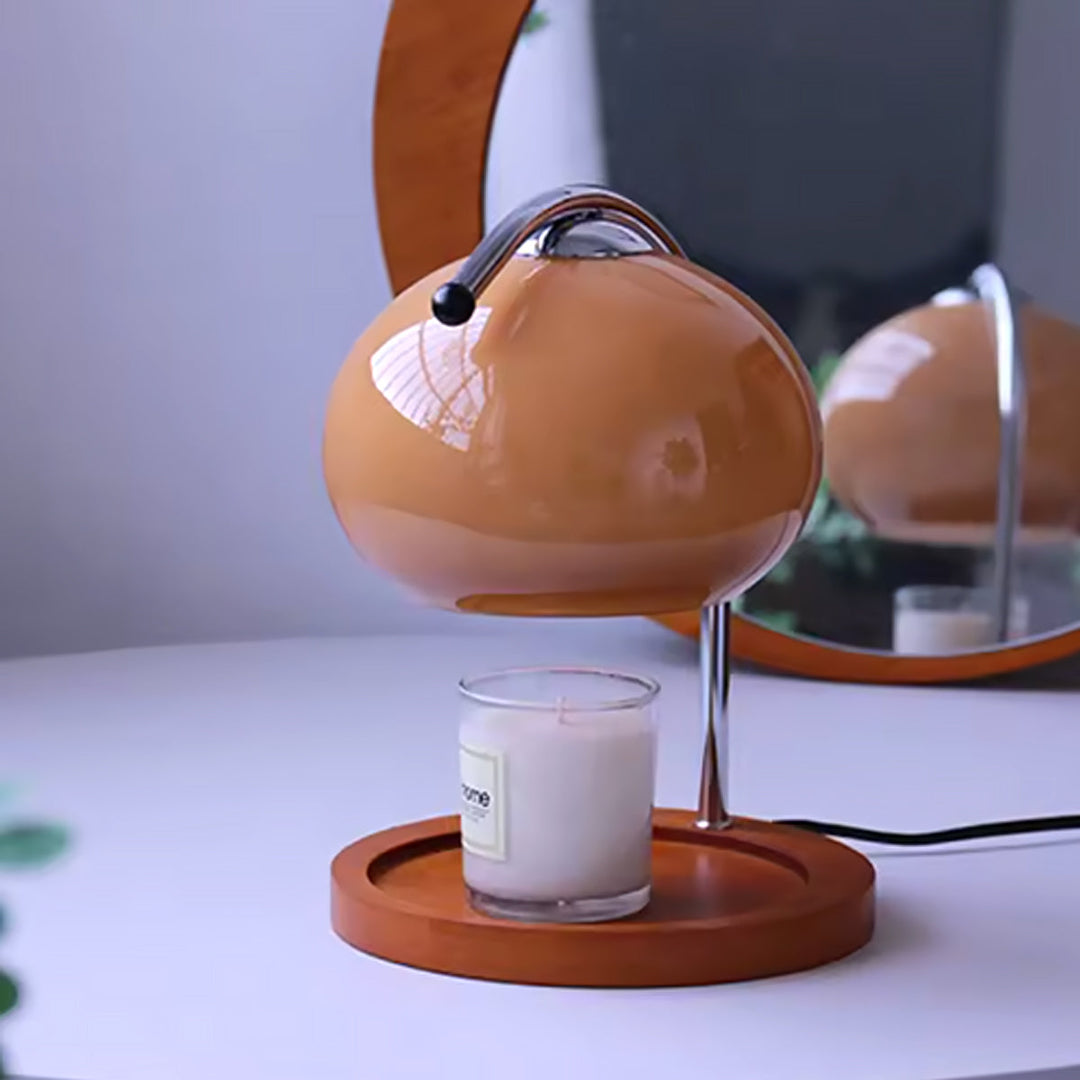 A glass candle sits on a wooden base beneath a dimmable metal and glass candle warmer lamp.