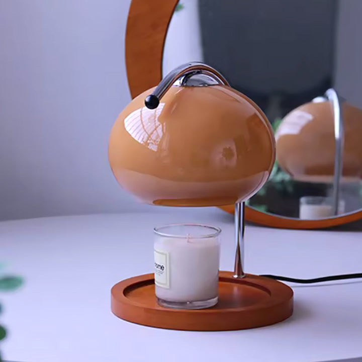 A glass candle sits on a wooden base beneath a dimmable metal and glass candle warmer lamp.