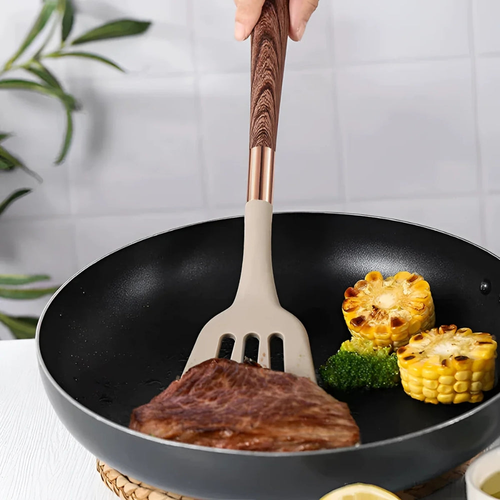 A silicone spatula with a wood-grain handle is used to flip a steak in a non-stick pan with corn and broccoli.