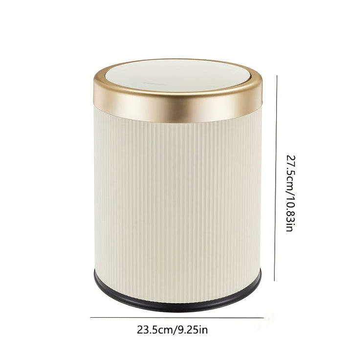 Cream oval trash can with a gold press-top lid and ribbed texture. Dimensions: 23.5cm/9.25in wide, 27.5cm/10.83in tall.