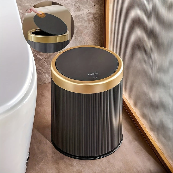 Black oval trash can with gold trim and a black press-top lid, next to a toilet.