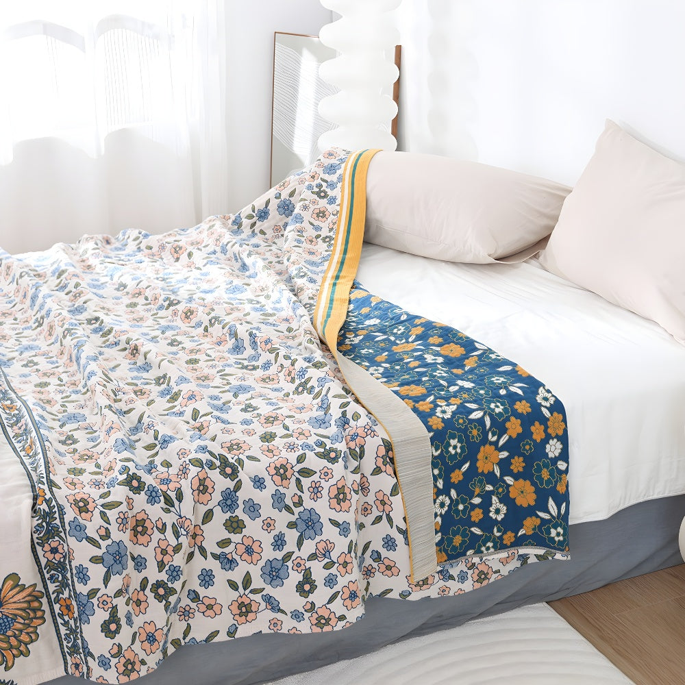 A boho floral blanket with a white base and blue and pink flowers, folded on a bed with pillows.