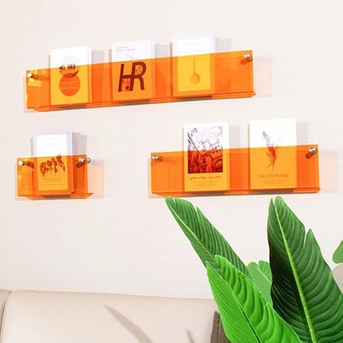 Three orange acrylic floating shelves mounted on a white wall, displaying books and cards.