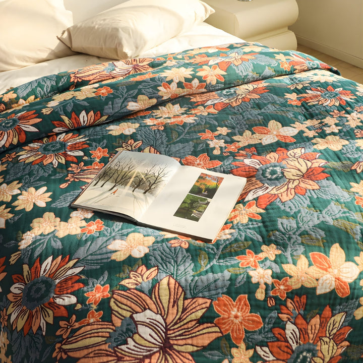 A floral patterned cotton throw blanket in green with orange and cream flowers, with an open book on top.