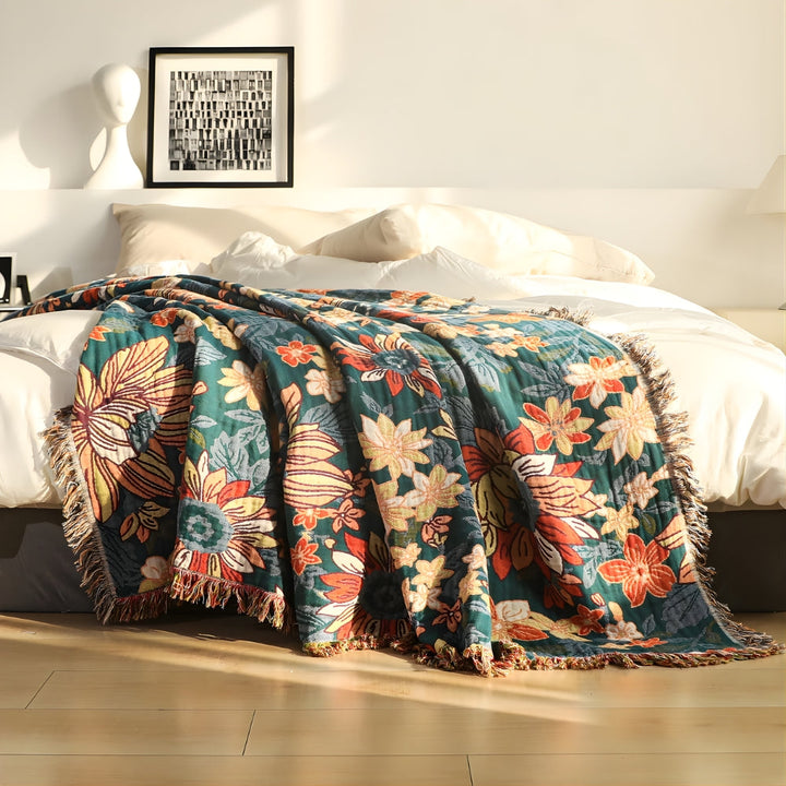 A teal cotton throw blanket with a large floral pattern in red, yellow, and orange, featuring fringed tassels.