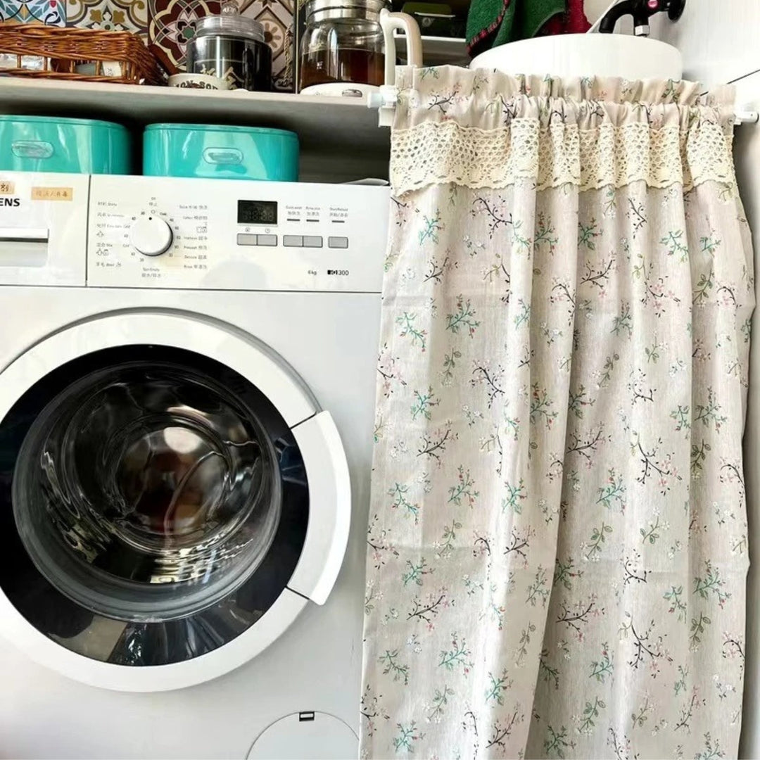 A light beige kitchen curtain with a delicate floral pattern of pink and green blossoms on branches.