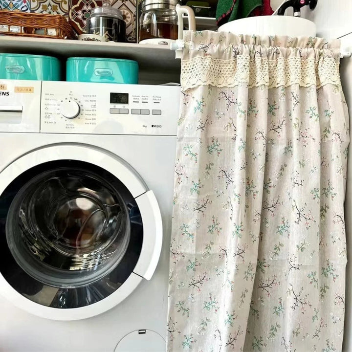 A light beige kitchen curtain with a delicate floral pattern of pink and green blossoms on branches.