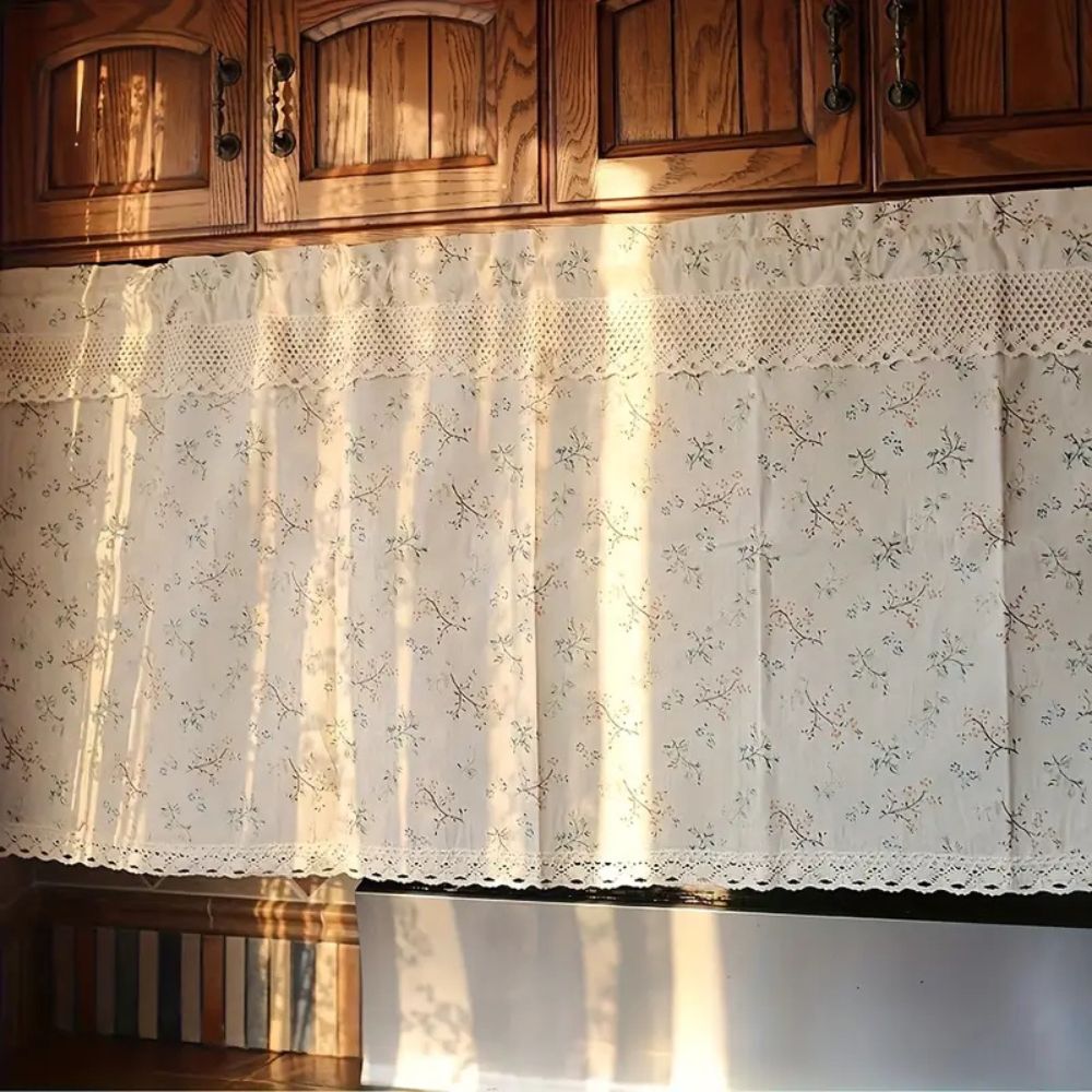White kitchen curtain with a delicate floral pattern and lace trim, hanging below wooden cabinets.