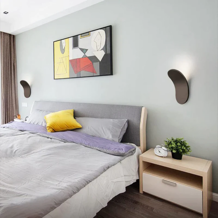 Minimalist LED wall sconce with a dark curved design mounted on a light blue wall above a bed.
