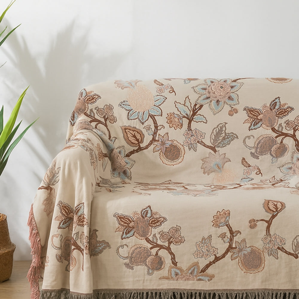 Cream colored cotton blanket with a floral trail pattern in brown, blue, and pink, featuring tassels on the edges.