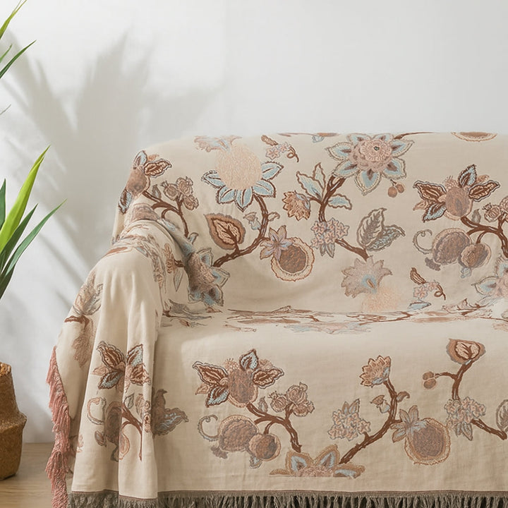 Cream colored cotton blanket with a floral trail pattern in brown, blue, and pink, featuring tassels on the edges.