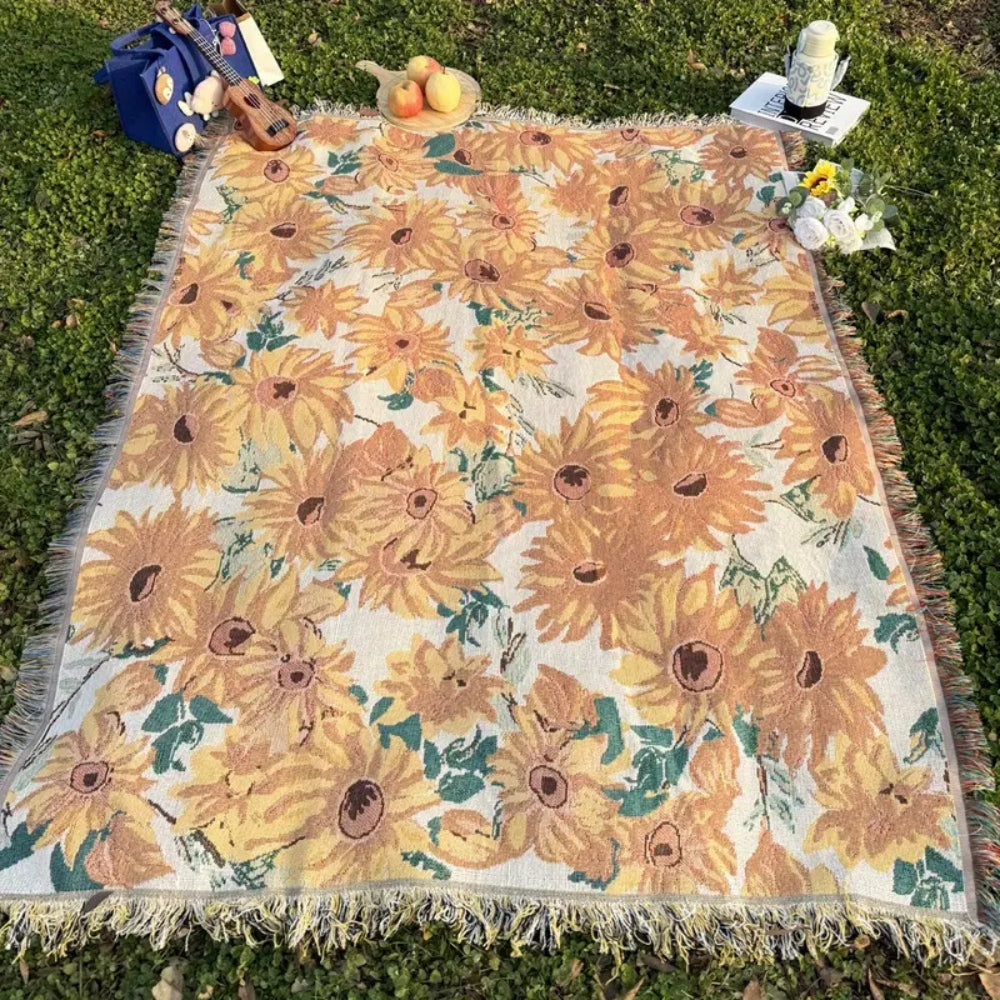 Floral picnic blanket with sunflowers and green leaves, fringed edges, on green grass.