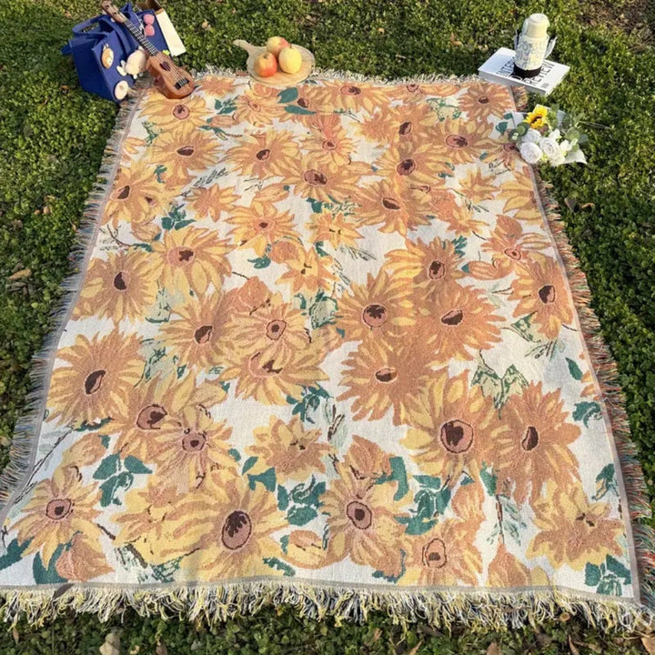Floral picnic blanket with sunflowers and green leaves, fringed edges, on green grass.