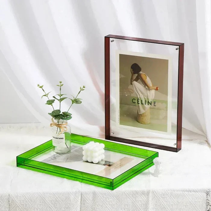 A transparent acrylic picture frame with a dark brown border stands behind a green acrylic tray holding a small plant and a white cube candle.